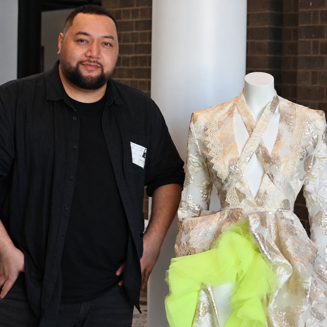 Iki Haangana in a black shirt standing beside a cream brocade gown with ruffled lime tulle on a mannequin